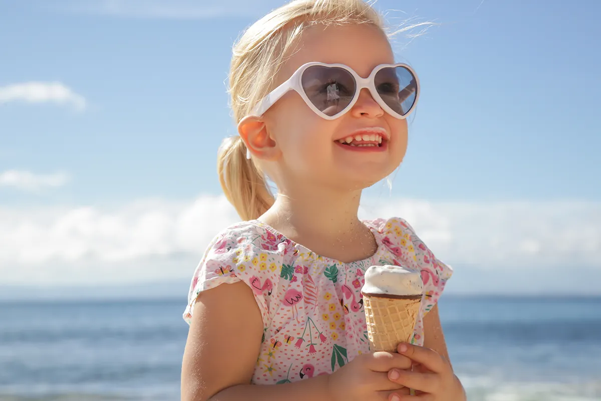 a little girl standing outside at the beach holding an ice cream cone and wearing sunglasses